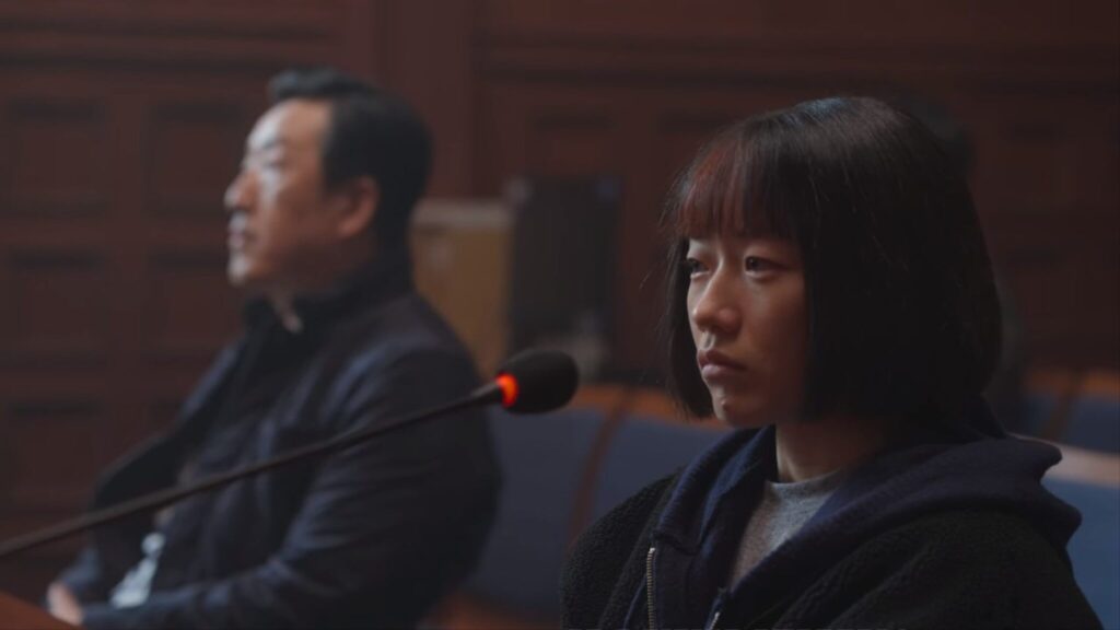 Jovem sentada em frente a um microfone dentro de um tribunal de justiça e ao fundo um homem sentado próximo a ela