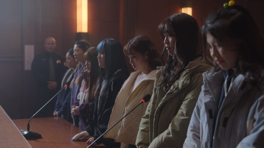 Sete jovens mulheres de pé em frente a uma bancada com microfones dentro de um tribunal de justiça com um policial ao fundo